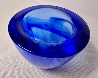 May include: A small, round bowl made of translucent cobalt blue glass. The glass features a swirling, marbled pattern of lighter and darker blue tones throughout its thick, smooth walls, creating a deep, vibrant appearance with a wide opening.
