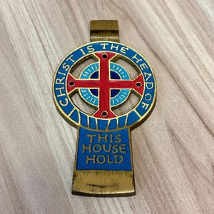May include: A gold-toned metal cross-shaped emblem with the text "CHRIST IS THE HEAD OF" in blue lettering around a red cross. The bottom section reads "THIS HOUSE HOLD" in blue. The emblem has a loop at the top.