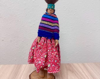 Könnte beinhalten: Eine Holzfigur auf einem runden Sockel mit einem braun geschnitzten Kopf, einem türkisfarbenen Stirnband und einer dunkelbraunen Kopfbedeckung. Die Figur trägt einen mehrfarbig gestreiften Schal über einem roten Rock mit weißem Blumenmuster.