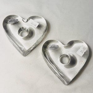 May include: Two clear glass heart-shaped candle holders. Each holder has a circular indentation for a candle and a small hole at the top. The holders have a textured surface and a sticker that reads "BLENKO HANDCRAFT".
