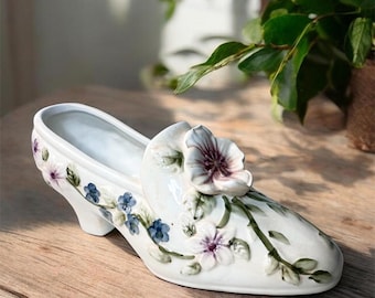 Könnte beinhalten: Ein weißes, schuhförmiges Dekorationsobjekt aus Keramik mit glänzender Oberfläche, verziert mit handgemalten blauen und violetten Blumenmotiven und grünen Ranken, gekrönt von einer großen, dreidimensionalen skulptierten Blume am Vorderblatt.