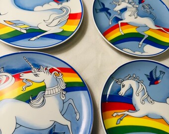 May include: Set of four decorative plates featuring a white unicorn with a rainbow and blue birds against a light blue background. Each plate has a rainbow border and a white rim. The plates are likely ceramic.