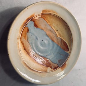 May include: A handmade ceramic plate with a unique design. The plate features a light blue, brown, and beige color scheme. The center of the plate has a light blue design, with brown and beige accents around the edges. The plate is round and has a slightly raised rim.