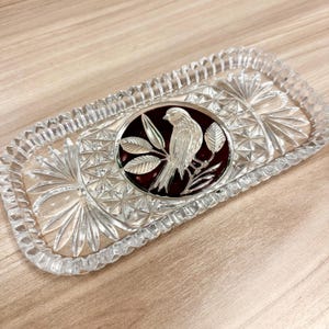 May include: A rectangular clear glass tray featuring a faceted, starburst-patterned border and a central circular medallion. The medallion contains a deep red background with a silver-toned relief of a bird perched on a leafy branch.