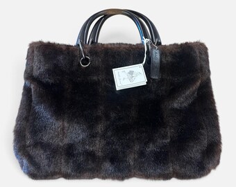 Könnte beinhalten: Eine dunkelbraune Kunstfell-Handtasche mit zwei abgerundeten, dunkelbraunen Holzgriffen, die an silberfarbenen Metallringen befestigt sind. Ein weißes Papieretikett mit einer kleinen Illustration und dem Text Isabelle Fiore hängt an einem Griff.