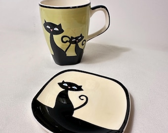 May include: A ceramic mug and a matching square saucer featuring stylized black cat illustrations. The mug has a light green body with two black cats, while the saucer is cream-colored with a single black cat. Both pieces have black rims.