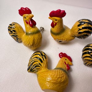May include: A set of three rooster-shaped cabinet knobs. Each knob is painted yellow with black-tipped tail feathers, a bright red comb, and red wattles. The roosters are depicted in a standing position with textured feather details and a metal screw.