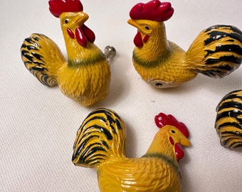 May include: A set of three rooster-shaped cabinet knobs. Each knob is painted yellow with black-tipped tail feathers, a bright red comb, and red wattles. The roosters are depicted in a standing position with textured feather details and a metal screw.