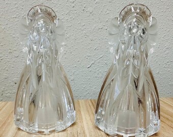 Könnte beinhalten: Zwei klare Glasengel-Figuren, die nebeneinander stehen. Jede Figur hat einen glatten, durchscheinenden Körper mit geformten Flügeln und einem Heiligenschein-Detail an der Oberseite. Das Glas ist reflektierend und zeigt klare Konturen.