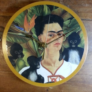 May include: Round wall clock featuring a portrait of Frida Kahlo surrounded by monkeys and tropical foliage. The clock has a gold-colored border and gold hands. The background includes a bird of paradise flower.