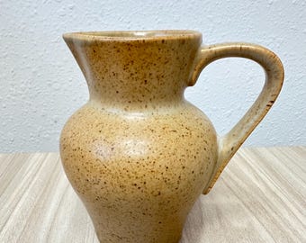 Könnte beinhalten: Ein Keramikkrug mit bauchigem Körper und geschwungenem Henkel, überzogen mit einer hellbraunen Glasur und einem dichten Muster aus dunkelbraunen Sprenkeln auf der gesamten Oberfläche, inklusive Ausguss und konischem Fuß.