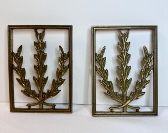 Könnte beinhalten: Zwei Vintage-Wanddekorationen aus Messing. Jedes Stück hat einen rechteckigen Rahmen, der ein detailliertes Messingblattdesign umschließt. Die Blätter sind symmetrisch innerhalb des Rahmens angeordnet und schaffen eine elegante und klassische Ästhetik. Das Messing hat eine warme, gealterte Patina.