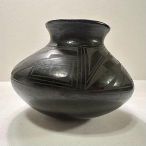 May include: A black ceramic vase with a rounded body and a flared rim. The vase features geometric designs in a darker shade of black, creating a contrasting pattern. The pottery has a smooth, glossy finish, reflecting the light.