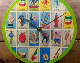 May include: A green and white clock with a round face. The clock face is decorated with images of playing cards from a traditional Mexican deck of cards called Loteria. The clock hands are black and point to the numbers 12 and 3.
