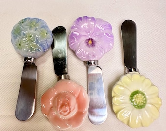 May include: Four metal spreaders with decorative ceramic handles shaped like flowers. The handles feature a blue hydrangea, a pink rose, a purple pansy, and a yellow daisy. Each spreader has a flat, silver-toned stainless steel blade.