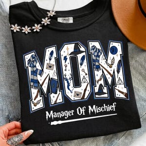 May include: A black t-shirt featuring the word MOM in large white block letters filled with blue and brown wizard-themed illustrations including scarves, hats, wands, and letters. Below the word, the text reads Manager Of Mischief.