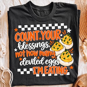 May include: A black t-shirt featuring a white and orange graphic with the text "COUNT YOUR blessings, not how many deviled eggs I'M EATING". The design includes a checkered border, small orange stars, and two illustrations of deviled eggs.