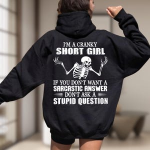 Może przedstawiać: Czarna bluza z kapturem z białą grafiką na plecach. Wzór przedstawia szkielet z podniesionymi ramionami oraz napis: I'M A CRANKY SHORT GIRL IF YOU DON'T WANT A SARCASTIC ANSWER DON'T ASK A STUPID QUESTION pogrubionymi białymi literami.