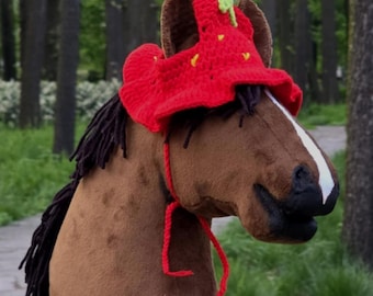 May include: A brown plush horse head with a red and green crocheted strawberry hat. The horse head is on a stick and is in a forest setting.