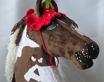 May include: A brown and white spotted horse head with a red crocheted strawberry hat. The hat has green leaves and a white crocheted ruffle.