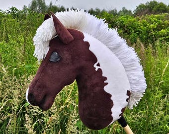 May include: A plush hobby horse with a brown and white head and a white mane. The toy horse has a black eye and is attached to a wooden stick. The background is a field of green grass and trees.