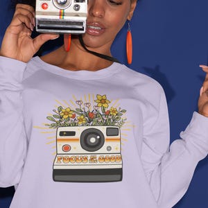Könnte beinhalten: Ein lavendelfarbenes Sweatshirt mit einer Vintage-Kamera-Grafik. Die Kamera ist mit Blumen gefüllt und hat den Text "FOCUS on the GOOD" aufgedruckt.