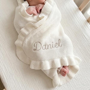 May include: Cream-colored baby blanket with a ruffled edge and the name "Daniel" embroidered in a light brown thread. The blanket is wrapped around a baby, with the baby's feet visible.