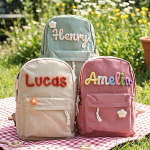 Puede incluir: Tres mochilas de pana en verde claro, beige y rosa, cada una con un nombre bordado en la parte delantera: "Henry", "Lucas" y "Amelia". Las mochilas tienen un bolsillo delantero y un adorno floral. Están sobre una manta a cuadros rojos y blancos.