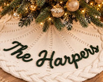 May include: A round, cream-colored knit Christmas tree skirt featuring a thick braided cable border and the dark green embroidered text "The Harpers" centered on the fabric. The skirt is positioned beneath a decorated evergreen tree.