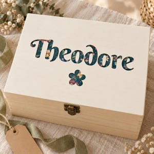 May include: A rectangular light-colored wooden keepsake box with a metal latch. The lid features the name Theodore in a cutout font, revealing a dark teal background with a colorful floral pattern, accompanied by a small flower-shaped cutout.