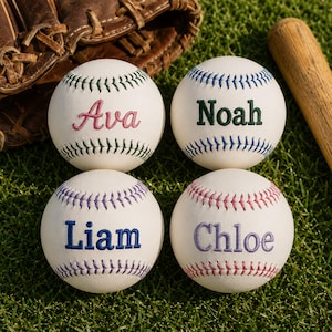 Op de afbeelding: Vier witte honkballen met gepersonaliseerde namen in verschillende kleuren: Ava (roze), Noah (groen), Liam (blauw) en Chloe (paars). Op groen gras, met een honkbalhandschoen en een knuppel op de achtergrond.