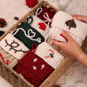Op de afbeelding: Een rieten mand gevuld met diverse gebreide kerstsokken. De sokken zijn in tinten rood, groen en crème, met feestelijke ontwerpen zoals kerstverlichting, harten en sneeuwvlokken. Eén sok wordt omhoog gehouden.