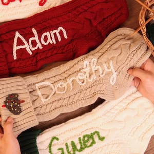 May include: Close-up of personalized Christmas stockings in red, cream, and beige. The stockings are knitted with cable knit patterns and feature names embroidered in white. A basket of yarn and knitting needles is in the background.