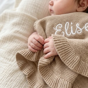 May include: A tan knitted baby blanket featuring a ribbed border and the name Elise embroidered in white cursive script. The fabric has a textured, woven appearance, and the blanket is draped over a small child, showcasing the soft knit construction.