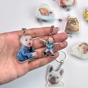 May include: Clear acrylic keychains featuring custom photos of babies, a child, a cat, and a dog. The keychains have silver-colored metal chains and rings. The baby and child keychains are held in a hand.