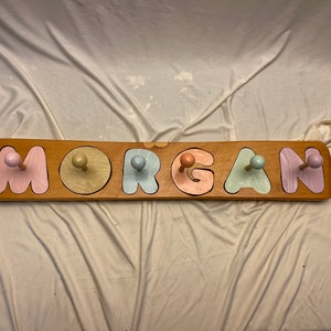 May include: Wooden name puzzle with the name "MORGAN" spelled out in colourful letters. Each letter has a knob on top, painted in pastel colours: pink, yellow, blue, pink, and purple.