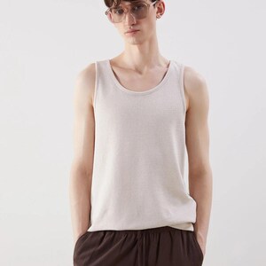 May include: A light beige tank top with a textured knit pattern. The tank top has a scoop neckline and is paired with brown pants. The person in the image is wearing clear-framed glasses.