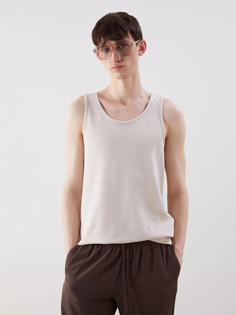 May include: A light beige tank top with a textured knit pattern. The tank top has a scoop neckline and is paired with brown pants. The person in the image is wearing clear-framed glasses.