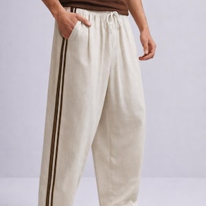 Puede incluir: Pantalones de lino color crema con cintura con cord&oacute;n y dos rayas marr&oacute;n oscuro en el lateral. Los pantalones de pernera ancha se combinan con zapatillas blancas.