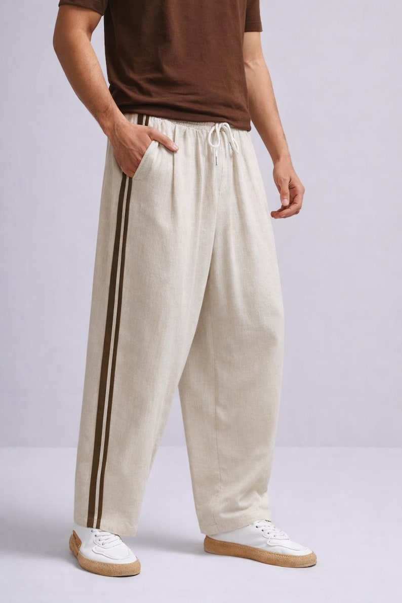 Puede incluir: Pantalones de lino color crema con cintura con cord&oacute;n y dos rayas marr&oacute;n oscuro en el lateral. Los pantalones de pernera ancha se combinan con zapatillas blancas.