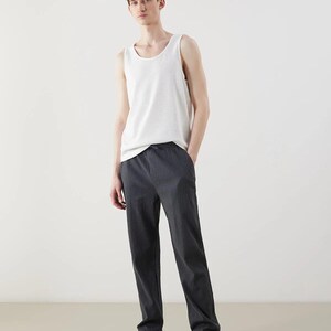 May include: A white tank top and dark gray trousers. The tank top is sleeveless and has a rounded neckline. The trousers are straight-legged and have a subtle vertical stripe pattern. White sneakers complete the outfit.