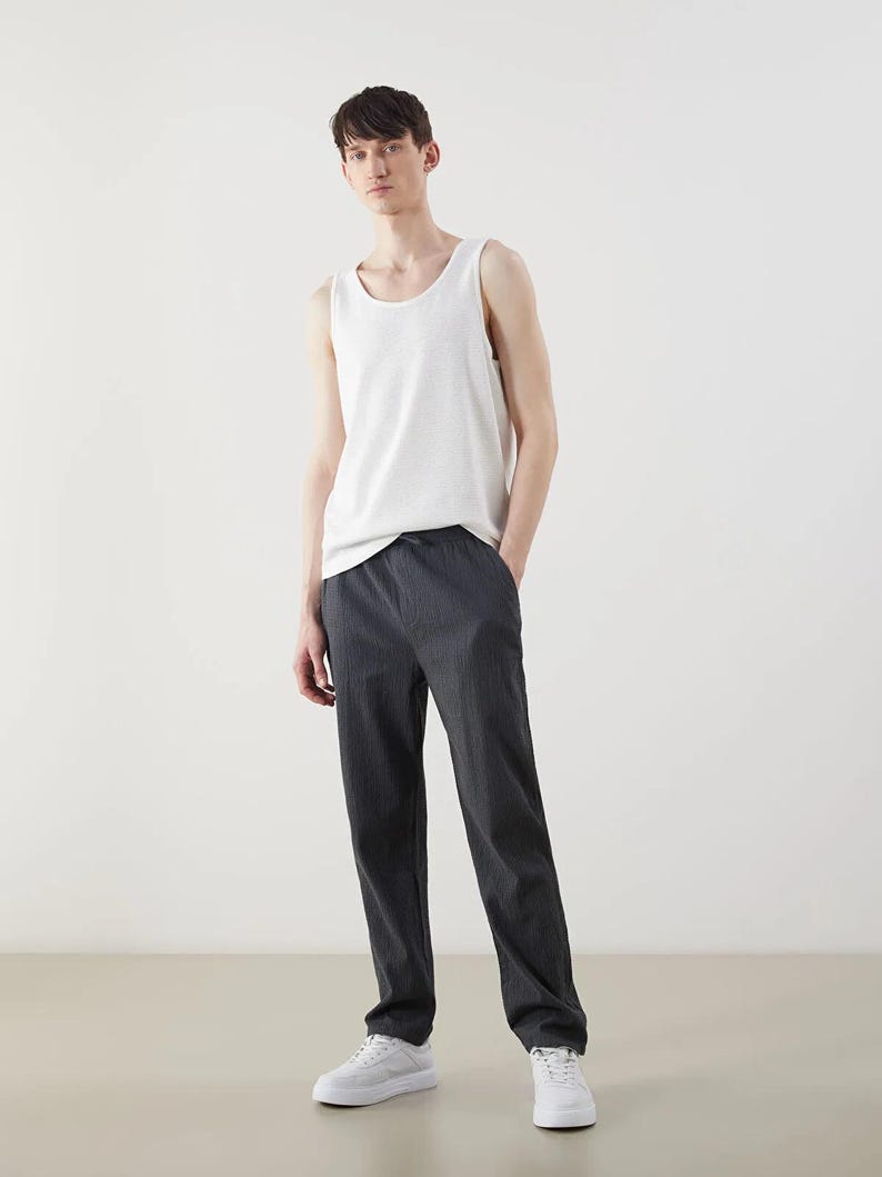 May include: A white tank top and dark gray trousers. The tank top is sleeveless and has a rounded neckline. The trousers are straight-legged and have a subtle vertical stripe pattern. White sneakers complete the outfit.