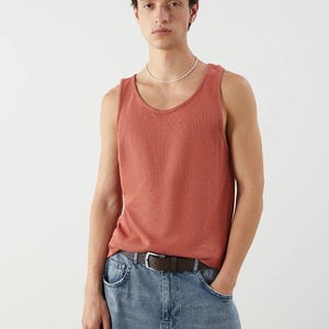 May include: A coral-colored tank top with a textured knit pattern. The tank top is paired with light blue denim jeans and a brown leather belt with a silver buckle. The model is wearing a white beaded necklace.