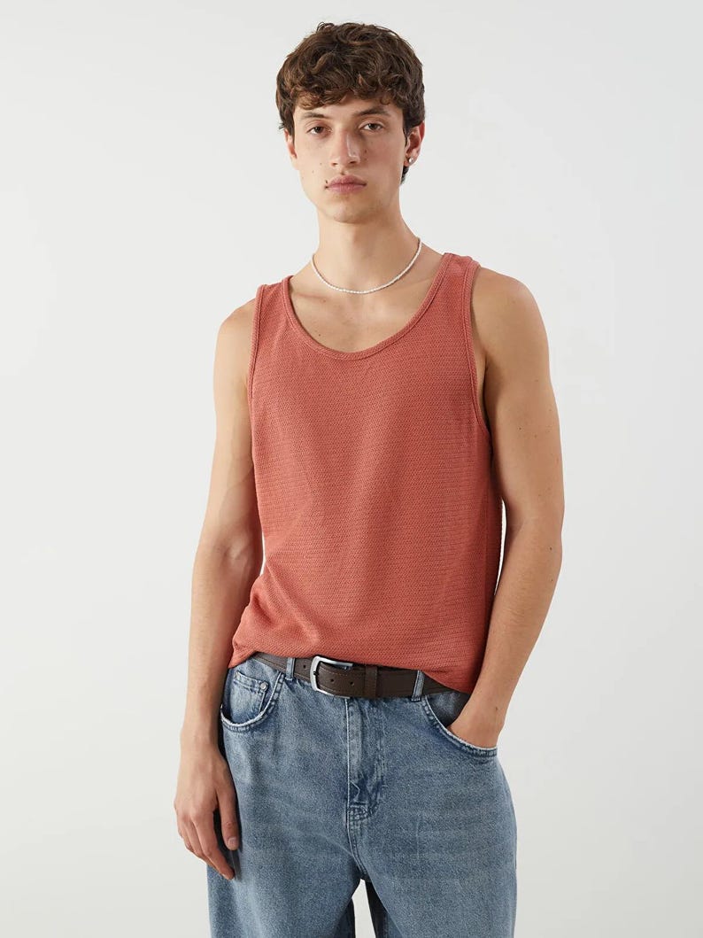 May include: A coral-colored tank top with a textured knit pattern. The tank top is paired with light blue denim jeans and a brown leather belt with a silver buckle. The model is wearing a white beaded necklace.