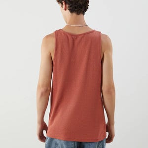 May include: A brick-red tank top with a textured knit pattern. The sleeveless shirt has a scoop neck and a straight hemline. The model is wearing blue denim jeans. The tank top is a casual, everyday garment.