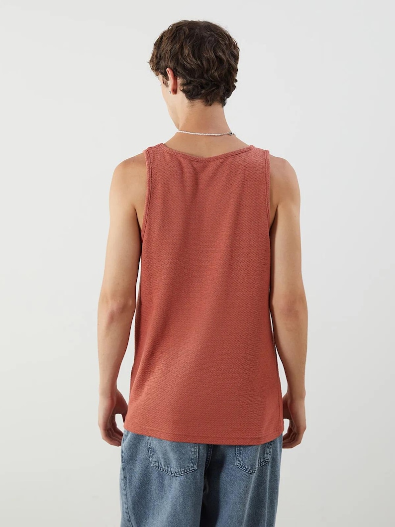 May include: A brick-red tank top with a textured knit pattern. The sleeveless shirt has a scoop neck and a straight hemline. The model is wearing blue denim jeans. The tank top is a casual, everyday garment.