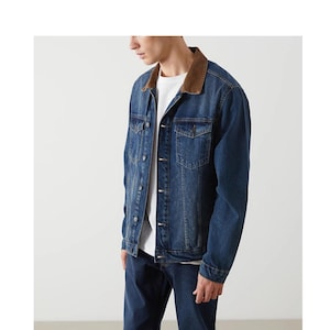 May include: A blue denim jacket with a brown corduroy collar. The jacket has two chest pockets and a button-up front. The person in the image is wearing a white t-shirt underneath and blue jeans.