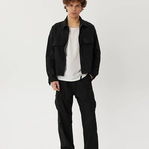 May include: A black utility jacket with a zipper closure and two flap pockets. The jacket is worn over a white t-shirt and black cargo pants.