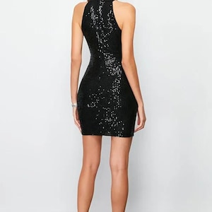 May include: A black sequin mini dress with a high neckline. The dress is sleeveless and form-fitting, with a subtle sheen. The model is wearing black high heels.