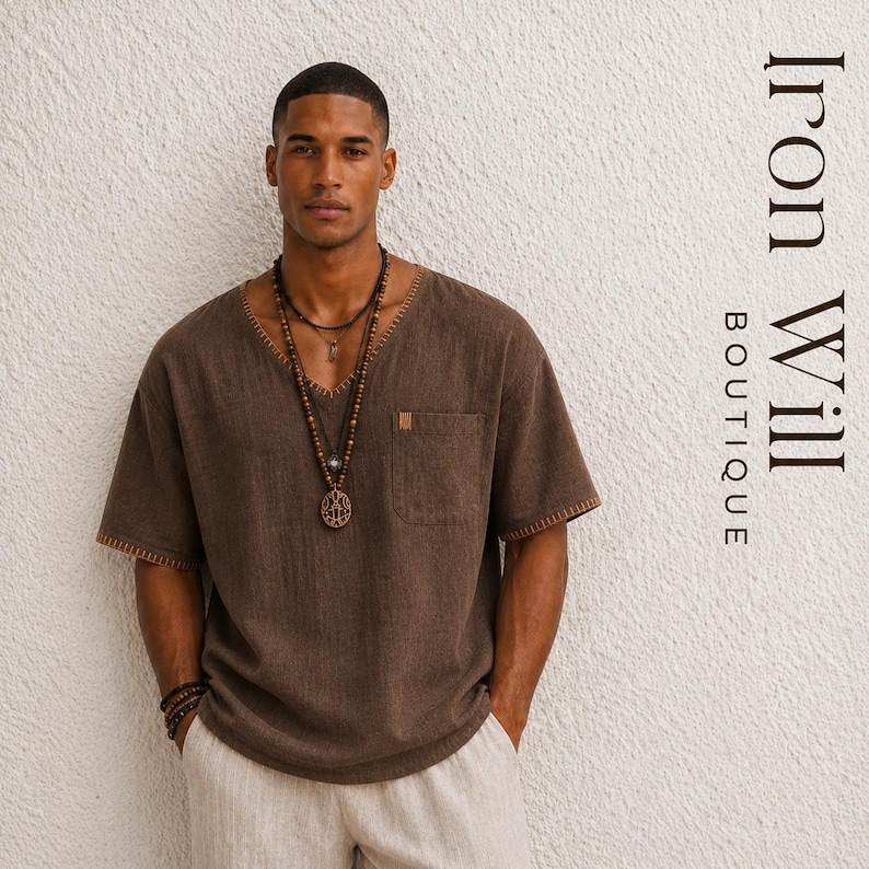 May include: A brown short-sleeve shirt with a V-neck and a pocket on the chest. The shirt has decorative stitching around the neckline and sleeves. The image also shows a wooden beaded necklace and the text "Iron Will Boutique".